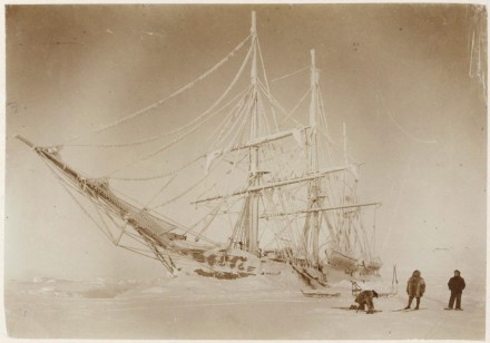 A black and white photo of the ship stuck in the Antarctic ice, with three crewmembers on the foreground.