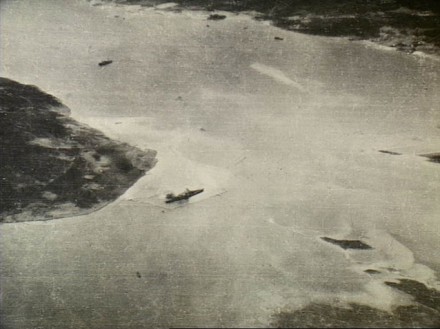 A black and white areal photograph showing the ship at anchor near an island.